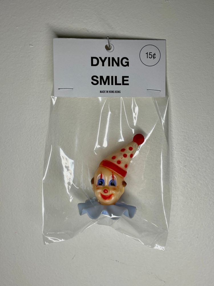 Photo of a Tozzi Toys bagged toy hanging on a white background which shows a vintage plastic cake topper smiling clown head with red and white polka dot hat and light blue neck ruffle collar. Clown head is packaged in a clear cellophane bag with a header card with printed text that reads “DYING SMILE” “Made In Hong Kong” and “15¢”