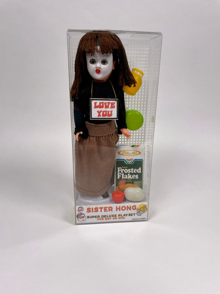 Photo of a custom-made Sister Hong doll playset in a clear plastic box. The small doll has a painted white face and red lips. It is wearing a drab brown skirt, black shirt, black shoes and a small sign around its neck reading “LOVE YOU” in red lettering. The playset includes a tiny green plastic pan, a yellow jug, an apple, potato and a box of Frosted Flakes. 