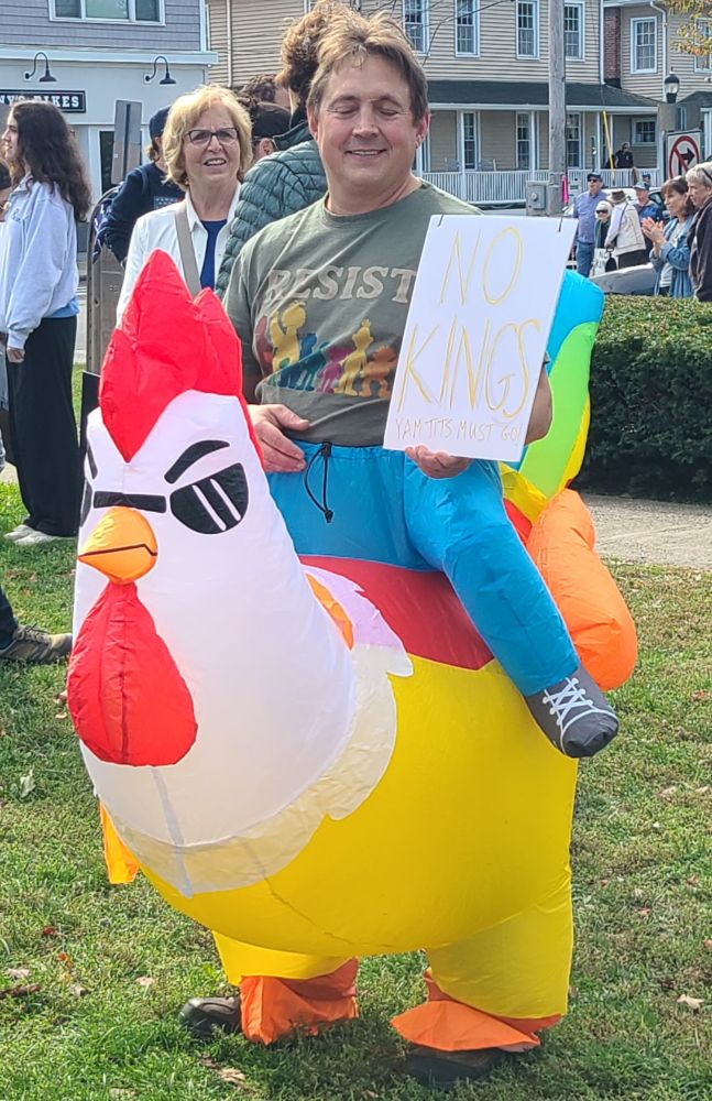 Man in an inflatable suit designed to make him look like he's riding a chicken, holding a protest sign: "No Kings - yam tits must go: