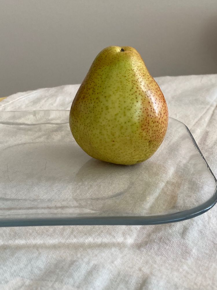 A green and reddish pear without a stem sits on a square, clear glass plate.