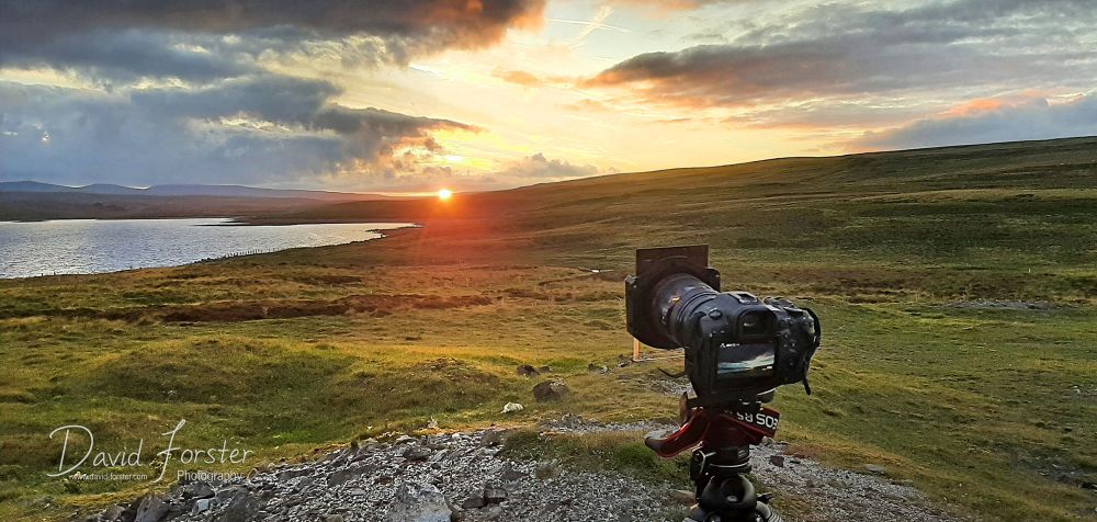 The sun sets over a remote unpopulated moorland with Cow Green reservoir and the high Pennine fells of Cross Fell and the Dun Fells on the horizon. 