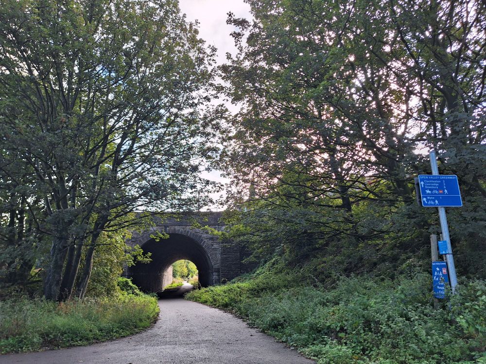 Photo shows a paved path under an old railway bridge with trees and hedges along the sides of the path. There is a pole with blue signs about the route and directions. 