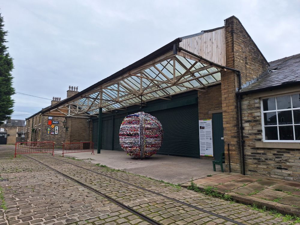 Photo shows a huge multicoloured ball of yarn under the stables canopy roof outside in the grounds of Bradford Industrial Museum. It is 3 metres tall and wide. The street is cobbled with tram tracks. 