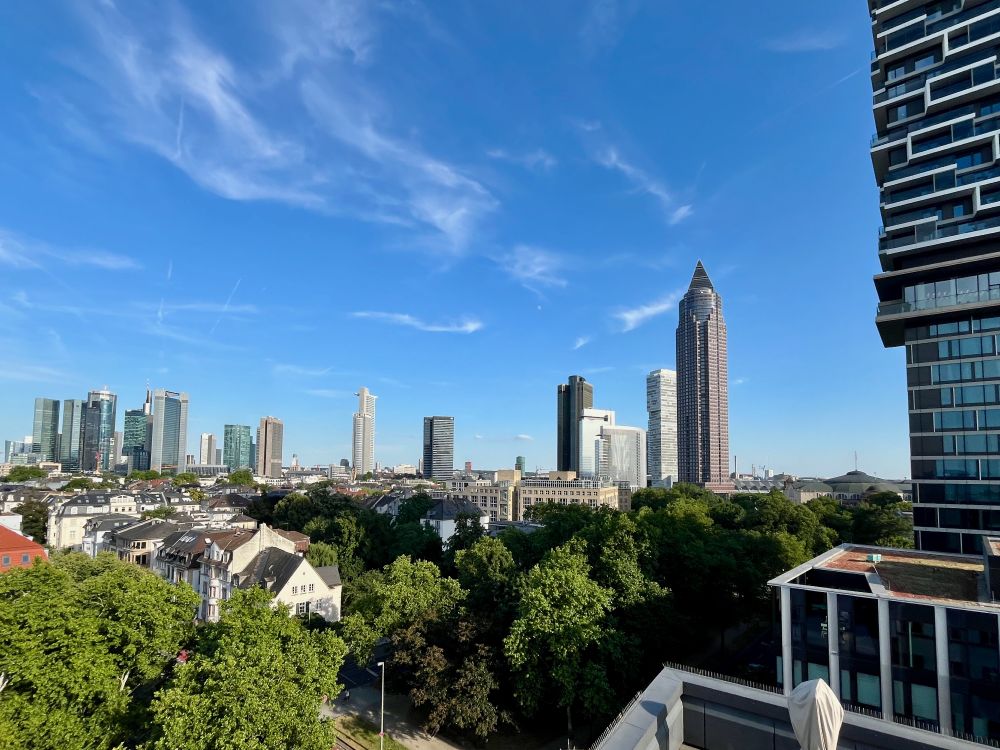 Skyline of Frankfurt am Main as seen from the rooftop observatory of the Physikalischer Verein Frankfurt, with trees in the foreground and skyscrapers in the back, in front of a blue sky.