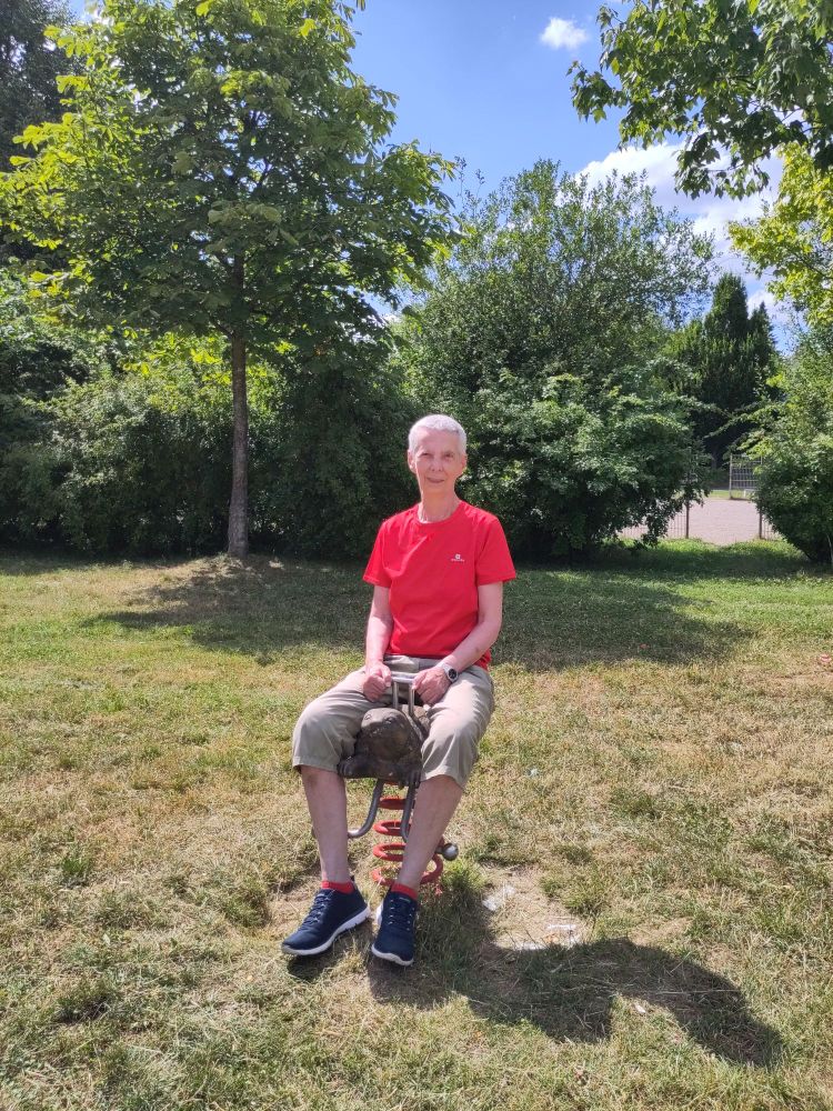 Ich sitze im Park am Spielplatz auf einer hölzernen Schaukelschildkröte und trage ein rotes T-Shirt, helle Bermudas, rote Sneakersocken und blaue Sneaker. 