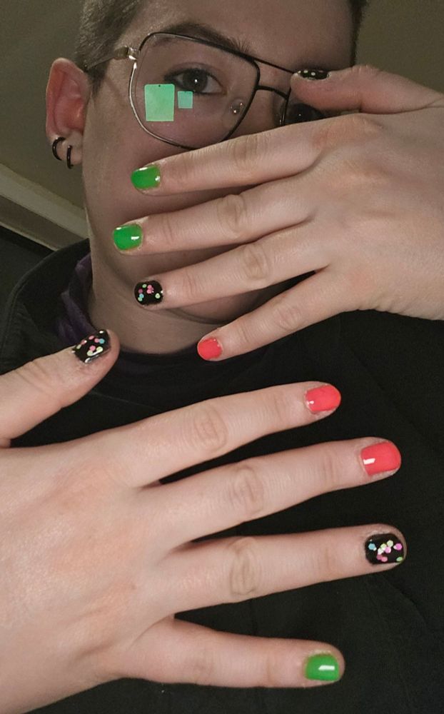 A picture of me, a white person, holding both hands in front of my face to show that they are alternatingly colored neon green, neon pink, and black with multicolored confetti nail polish.