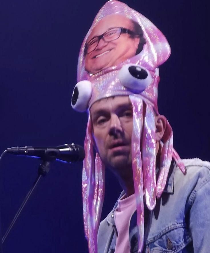 Blur & Gorillaz frontman Damon Albatn on stage, wearing a medusa-shaped hat with actor Danny DeVito's face on it. 