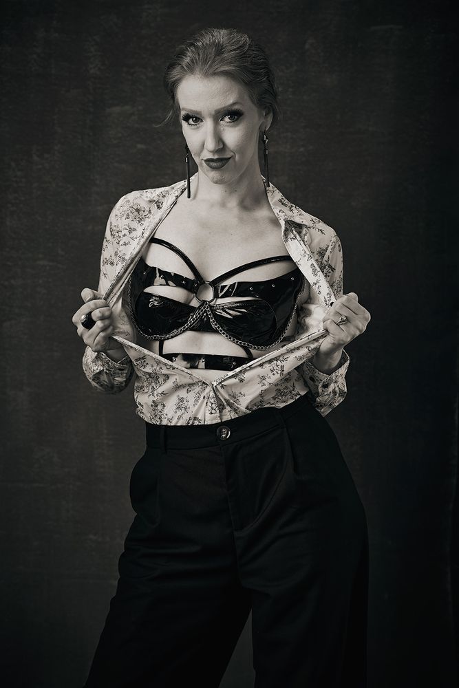Black and white photo of Sonia Harcourt pulling open her shirt to reveal the kinky bodice underneath, while making defiant eye contact with the viewer.