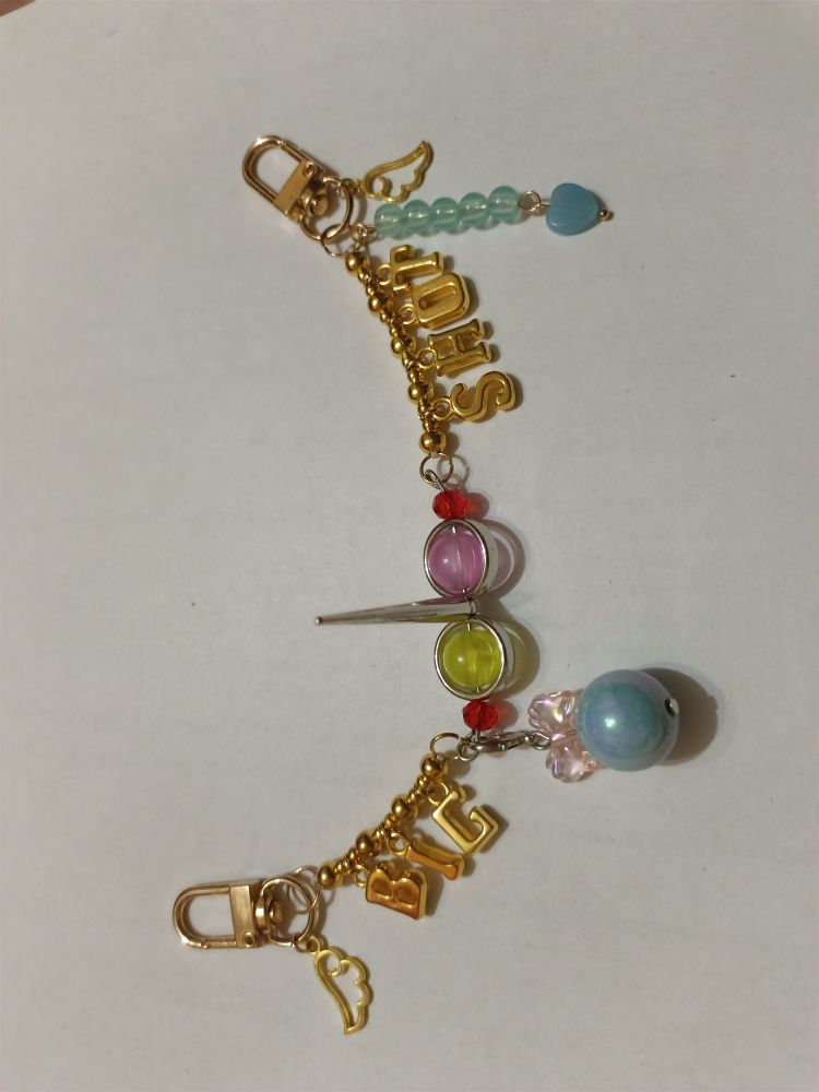 a long keychain with two carabibers on each side. it's themed after spamton g. spamton from deltarune, with a bead formation reminiscent of his face in the middle (two large beads for his eyes, a spike in the middle for his nose, and two smaller red beads at each side for his cheeks), and golden words "BIG" "SHOT" to the sides of it. there are also wing charms, a charm reminiscent of his heart attack, and a big blue bead with a bow that's supposed to be the pipis. 