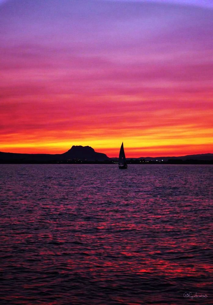 Photo au format vertical du rivage maritime à l’horizon, vu depuis la mer alors que le soleil vient de se coucher. Le ciel est couvert de nuages cotonneux qui ont pris des couleurs intenses, allant du parme en passant par le rose, le rouge, l’orange et le jaune. Presqu’au centre de l’horizon, on voit une formation rocheuse qui dépasse des plaines et se découpe en noir devant les nuages. À sa droite, on aperçoit un voilier en train de rentrer au port. La mer a pris une teinte violet foncé et les vaguelettes renvoient les reflets roses et orangés du coucher de soleil. 