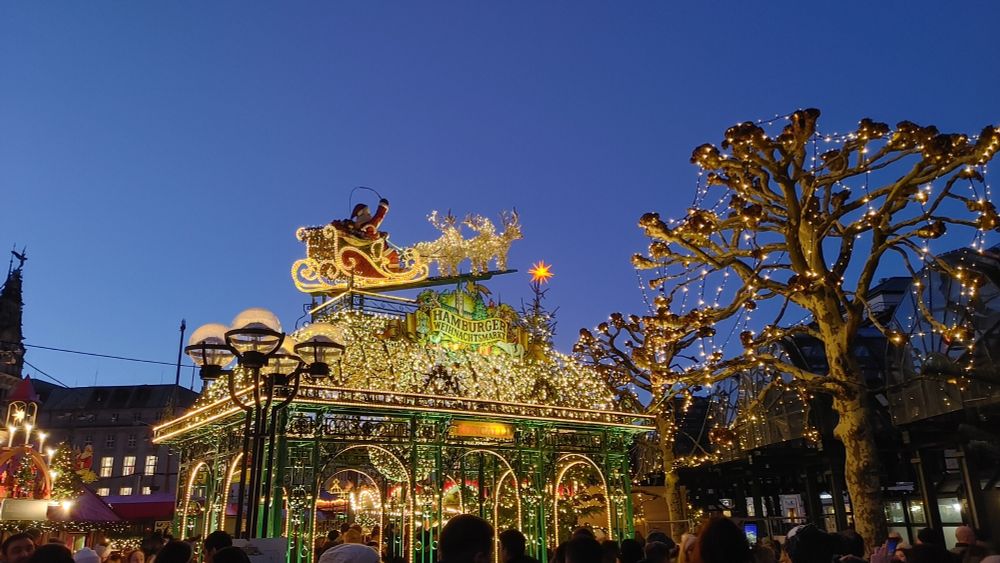 Hellbeleuchtetes in Gold und Grün.
Schlitten mit Rentieren auf einem Dach eines kleinem Gebäude aus Metallstreben.
Rechts ein Baum
Links und Rechts Häuser
