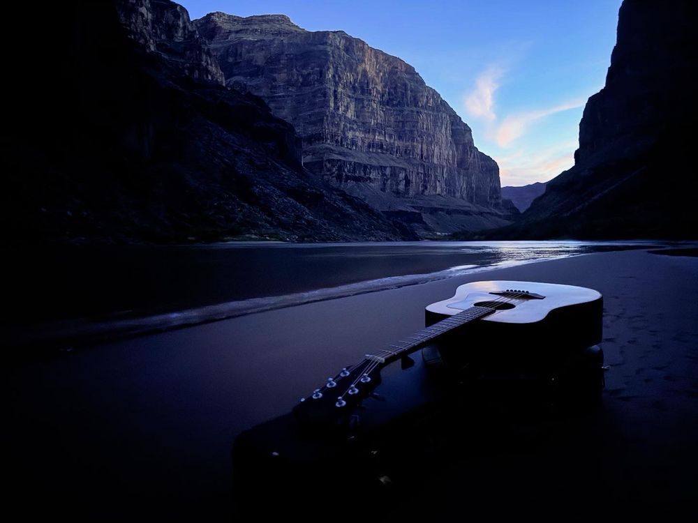 An acoustic guitar lays on a sandbar by the Colorado River running through the Grand Canyon reflecting the sunset’s fading light. 
