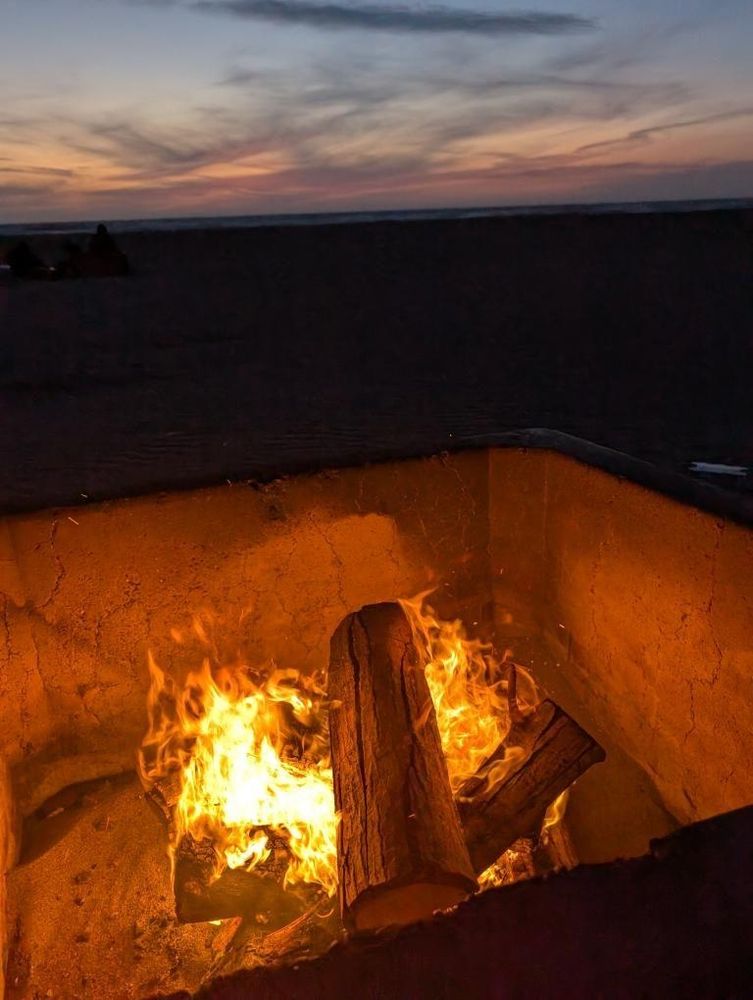 A small fire in a sheltered pit on the beach. The twilight sky with sparse clouds fills the background.