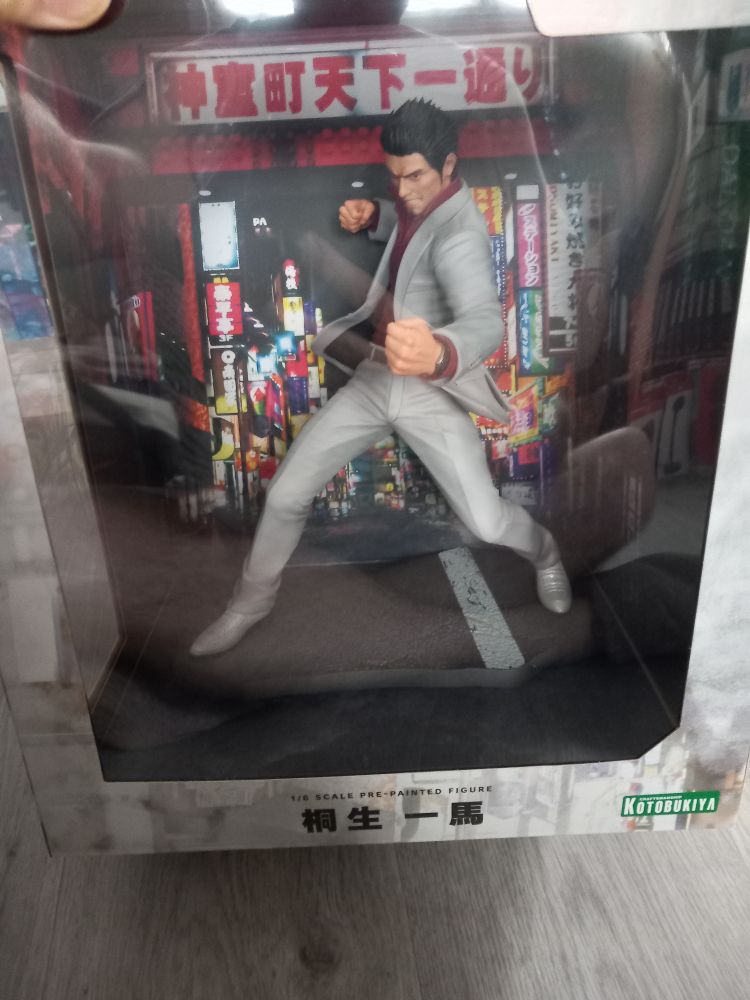 Figurine de Kazuma Kiryu, héros de la série Yakuza/Like a Dragon. Un homme aux cheveux bruns courts avec une chemise rouge et une veste et un pantalon gris clair et des chaussures de la même couleur.
Derrière la Figurine se trouve le décor principal de la série de jeu, Kamourocho, un quartier fictif de Tokyo inspiré de Kabukicho.