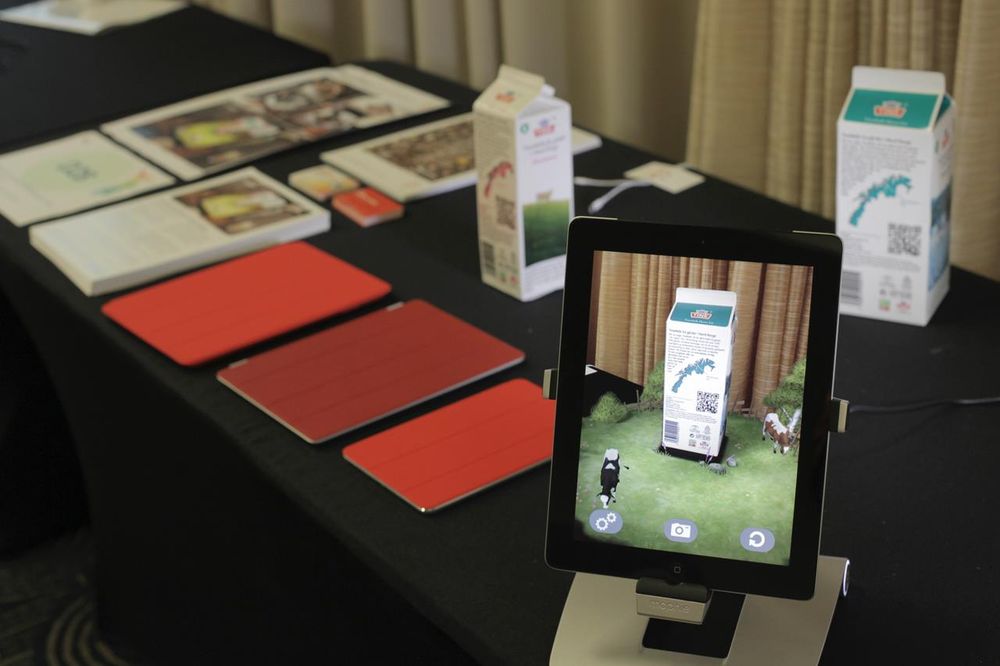 An iPad is seen on a stand on a table covered with a black cloth. The iPad screen show an AR app centered on a milk carton which is also on the table. We see two animated cows grazing on a small green field. On the table top are several other iPads with red covers (closed) and some stacked flyers/marketing material and another milk carton. 