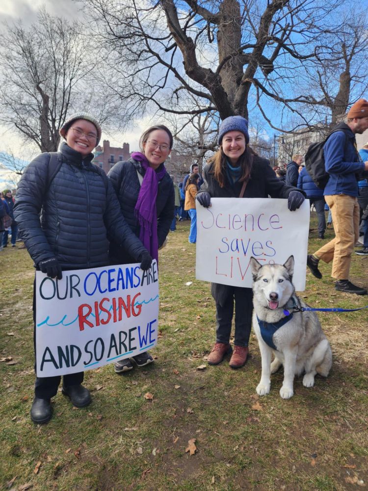 3 people and a dog. 2 people holding signs saying, "Our oceans are rising and so are we" and "Science saves lives."