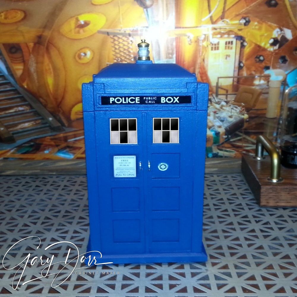 A handcrafted model of the TARDIS, designed as a blue British police box with the text 'POLICE PUBLIC CALL BOX' at the top. Placed on a patterned surface, the background mimics the detailed interior of the TARDIS. Signature 'Gary Dorr Cabinet Maker' displayed at the bottom left.
