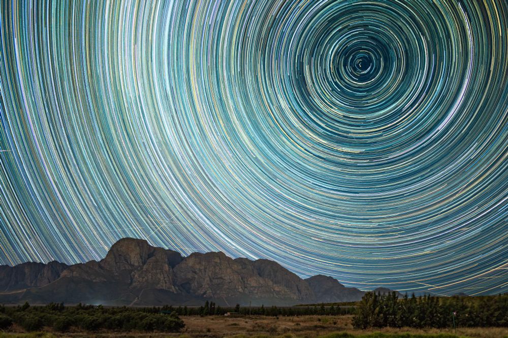 Star trails image captured on December 21, 2024, at Boschendal Wine Estate near Cape Town, oriented southward. The composite was created by stacking 600 individual 30-second exposures, illustrating the apparent rotation of stars around the southern celestial pole due to Earth’s rotation. Color variations in the trails correspond to stellar temperatures, with cooler stars appearing redder and hotter stars appearing bluer or white. Straight horizontal streaks across the frame are primarily satellites in stable orbits, accompanied by occasional bright meteors. The image shows minor camera movement artifacts resulting from windy conditions and the absence of a tripod. This technique visualizes Earth’s rotation.

Leica Q3 camera, 600 x 30s exposures, 28mm @ ƒ/1.7, ISO 100. Edited in Lightroom, stacked in StarStaX for Mac, final touches in Photoshop.