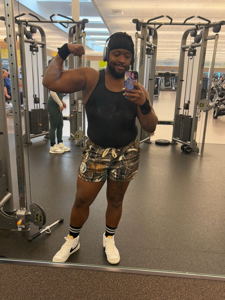 Picture of me flexing in the gym wearing a black tank top, camo shorts and white shoes.