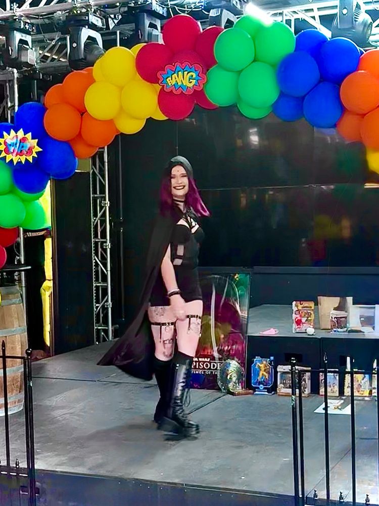 A person, dressed as Raven from Teen Titans, standing on a stage with prizes in the background. A balloon arch is overhead with various onomatopoeias from comics.