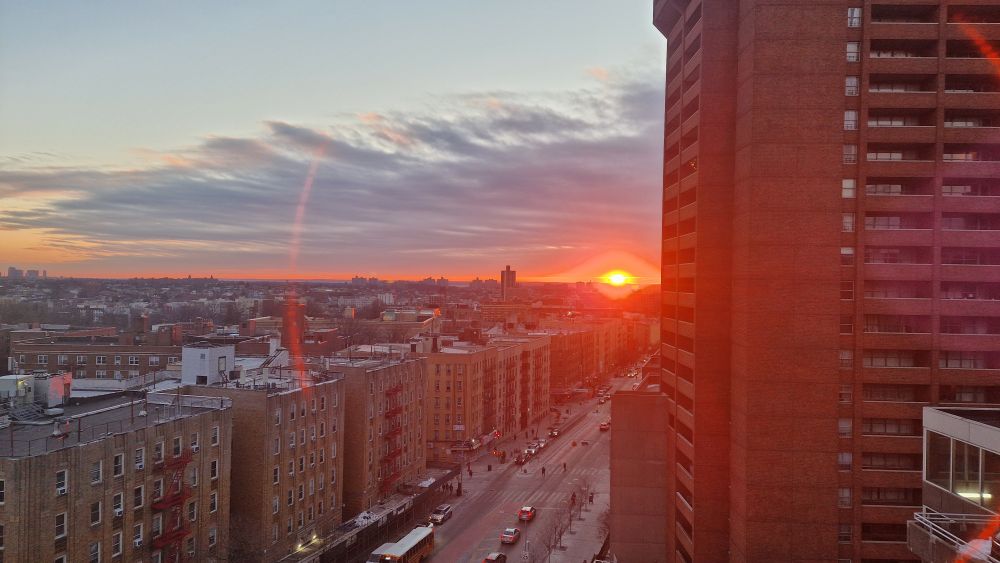 Ansicht eine Großstadt bei Sonnenaufgang in drei Bildanteilen. Rechts ein rot-braunes Hochhaus, das die ganze Bildhälfte einnimmt. Links unten die Gunhill Road in der Bronx, umsäumt von braunen Wohnblocks. Der Himmel (links oben) ist hellblau, leicht mit Wolken durchzogen und relativ zentral im Bild geht die Sonne als roter Glutball auf