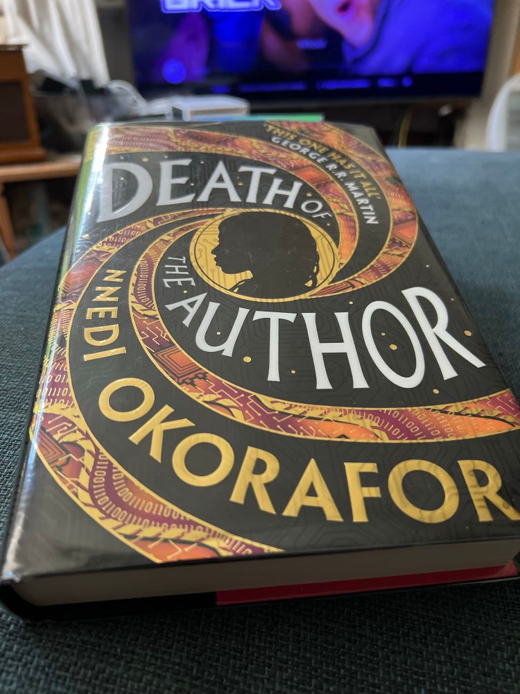 Library book copy of the hardcover book ‘Death of the Author’ by Nnedi Okorafor resting on a blue sofa. 