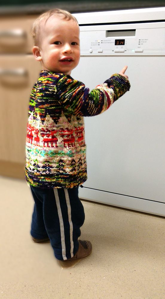 Photo of a baby standing unsteadily while wearing a multicolored handknitted cardigan with a reindeer motif. 