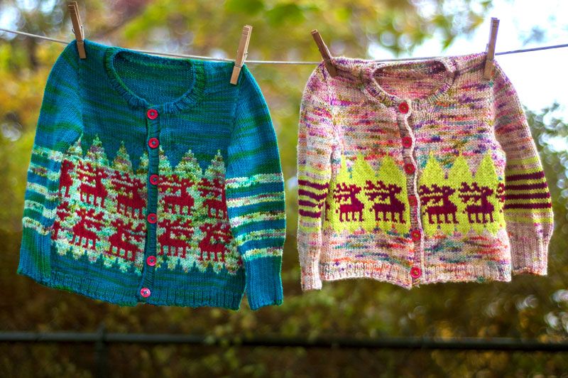 Photo of two handknitted baby cardigans hanging on a clothesline. One is mostly blue and green, the other mostly pink and neon yellow, but both have the same reindeer motif in red.