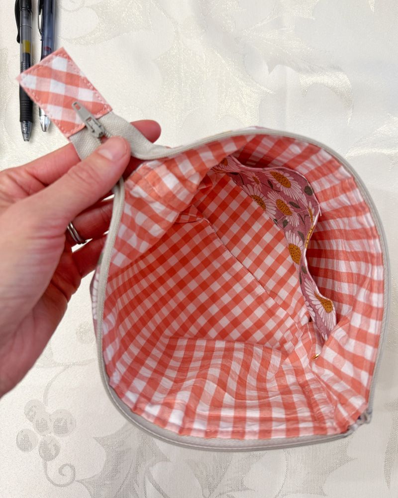 Inside of small zippered project bag with a coral-peach gingham interior fabric that matches the zipper tab, interior pockets that match the exterior mauve daisy fabric. 