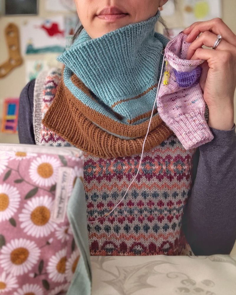 Stephanie, a tan skin woman, from smile through torso, sitting at a table, wearing a colorwork wool vest over a heathered dark purple-gray thin long-sleeve shirt and a blue and brown cowl covering her neck and upper chest. In hand is a speckled pink and purple sock in progress. 