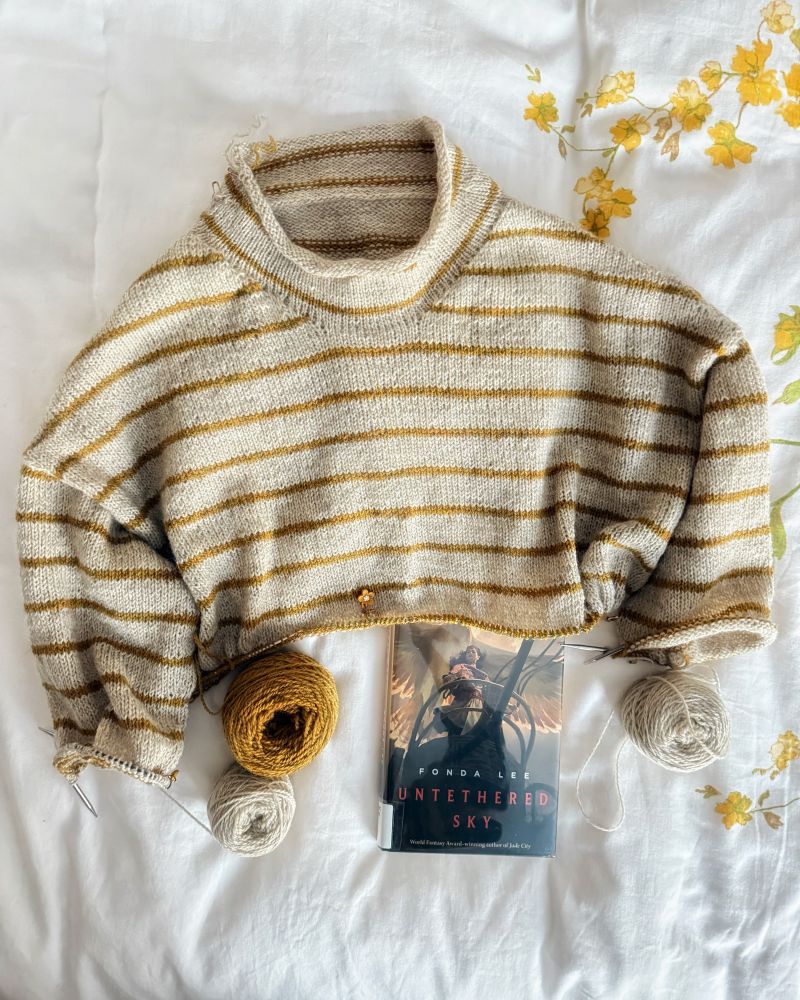 Drop-shoulder sweater in progress that’s a light gray-beige with heathered mustard yellow stripes throughout. It has a mock neck and live stitches on the body and the sleeves. Hardcover book features a young woman with dark hair standing on a chariot with an enormous bird flying above her.