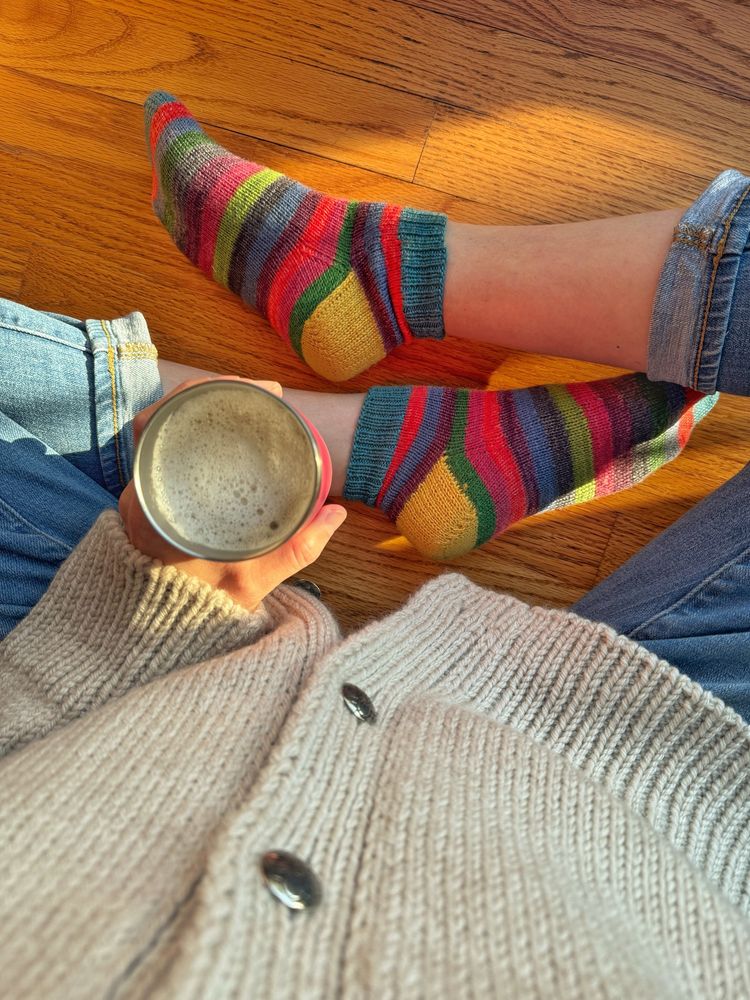 Angle looking down at Stephanie’s feet. She’s sitting on a hardwood floor. She’s wearing cuffed jeans, a beige-light gray wool jacket with pewter buttons. In hand is an insulated tumbler of hot coffee. On her feet are saturated striped wool socks morning sunshine. 