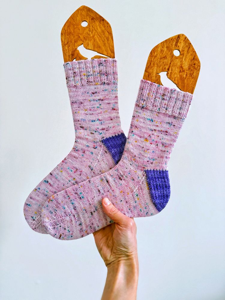 Completed handknit socks on bird-themed wooden sock blockers. Pink-lavender socks with multi-colored defined speckles and a purple slipper stitch heel flap and heel turn.