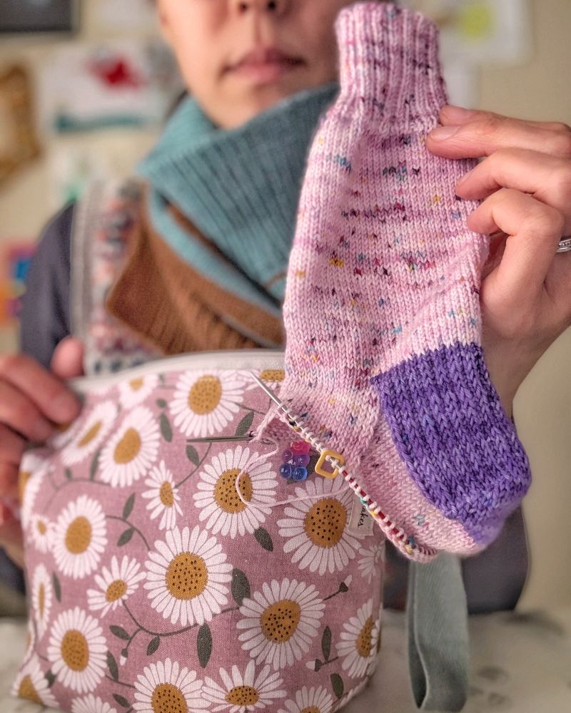 Close-up of sock project in hand - it’s knit cuff-down using a speckled pink and purple main yarn and a purple yarn for the heel. Live stitches on the foot. 