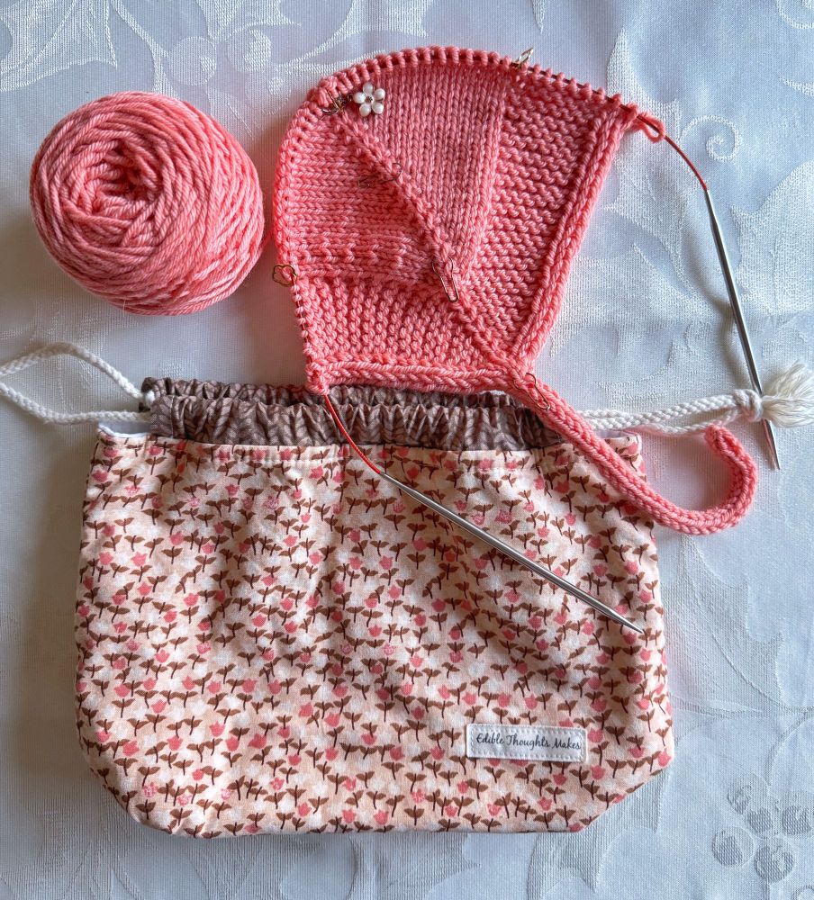 Bonnet-in-progress knit with a saturated bright peachy yarn. Garter texture and I-cord edging and tie. Floral drawstring project bag in brown, peach, cream.