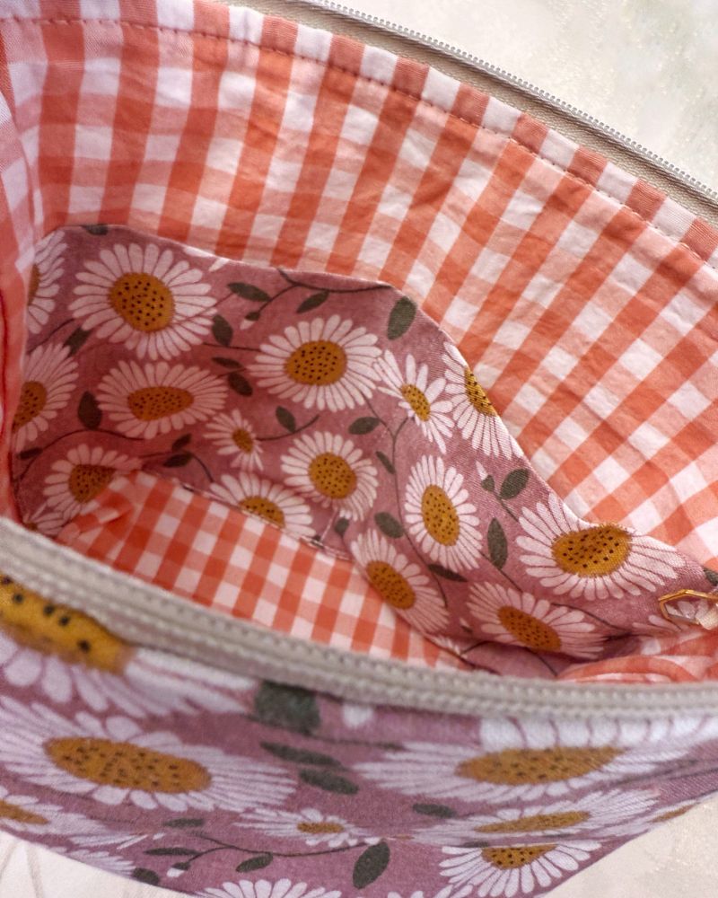 Small zippered project bag features a coral-peach gingham zipper interior fabric, interior pockets that match the exterior fabric that’s a mauve daisy print. 