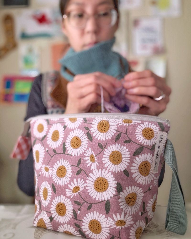 Focus on the small sock project bag featuring a mauve daisy print with a zipper top, coral-peach gingham zipper tab and a repurposed teal-gray side strap cotton loop handle. Behind Stephanie, out of focus, is a wall with a pair of wooden sock blockers and many drawings by her children.