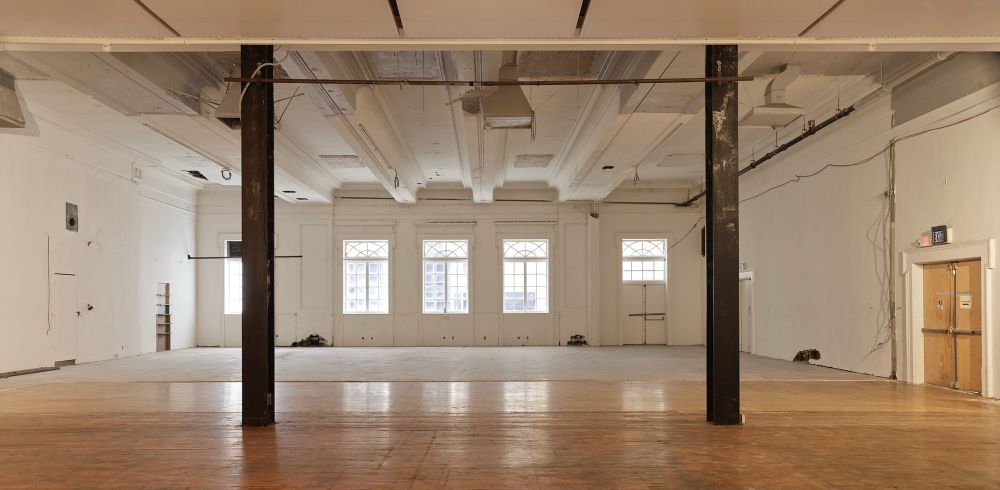 An image of two beams opening into a large hall with 17 foot ceilings. The wood flooring is covered by linoleum about halfway through the space. 