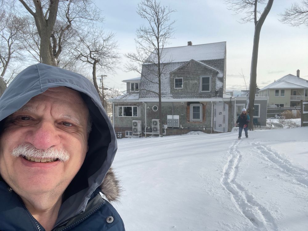 A man with a gray mustache and hood stands in a backyard blanketed by snow. 
