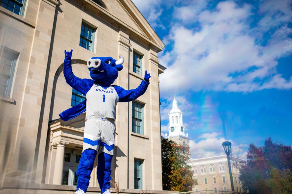 Victor E. Bull poses triumphantly on UB's south campus. He is wearing a cape. The clocktower of Hayes Hall can be seen in the background.