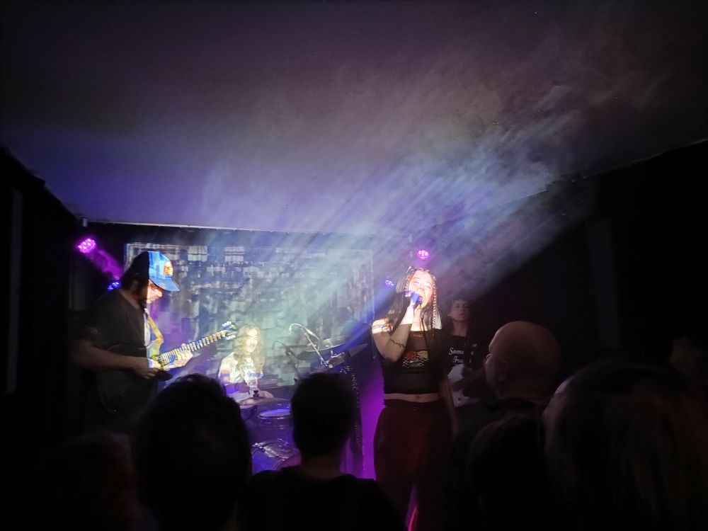 die band water from your eyes auf derselben bühne von psychedelischen projektionen angestrahlt, links der gitarrist mit mashbackkappe, im hintergrund der drummer, vorne die sängerin mit geschlossenen augen am mikro, rechts im halbdunkel die bassistin