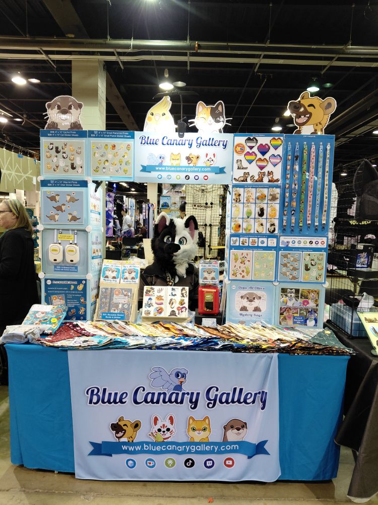 Blue Canary Gallery booth at MFF dealers den with solis the wolf as the merchant, half black and half white fursuiter