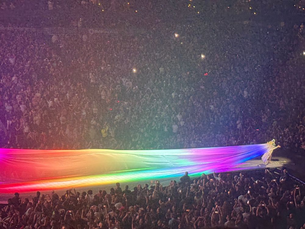 Lady Gaga stands at the edge of a runway stage in a packed arena, dramatically pulling an enormous flowing rainbow-colored fabric behind them. The fabric stretches across the stage, glowing with vivid hues under the lights, creating a striking visual that sweeps toward the cheering crowd. The audience is illuminated by scattered phone lights, capturing the magical, pride-filled moment. 