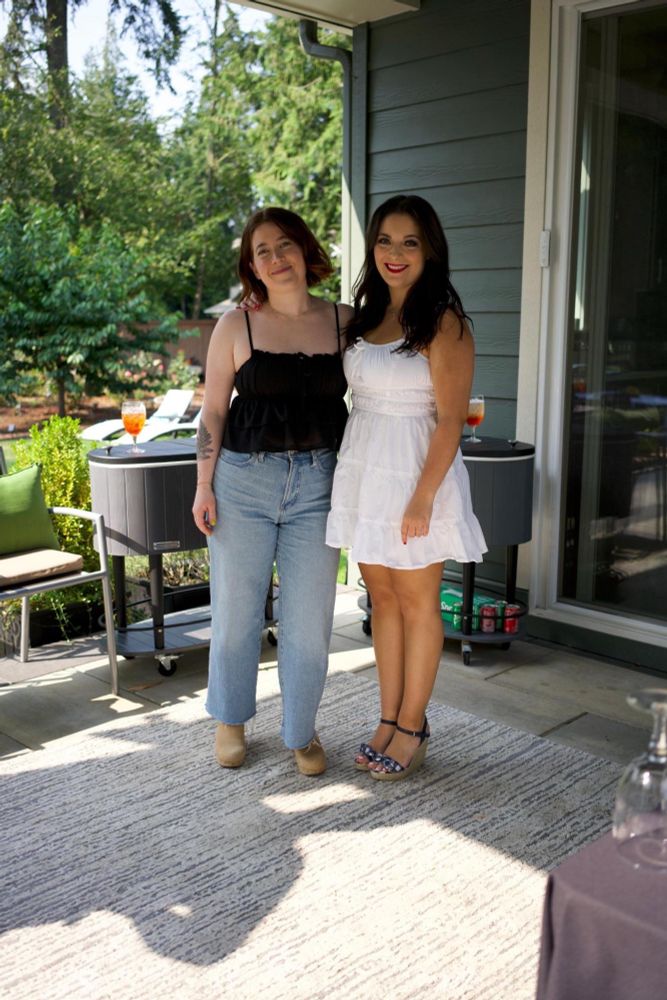Two women posing for a photo. Savannah is on the left. She has a brown bob and is wearing a blank spaghetti strap top, wide leg jeans and beige clogs. Claudia is on the right. She has long dark hair and is wearing a white sundress and wedges.