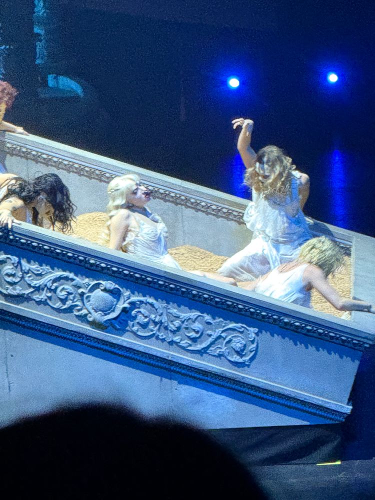 A group of performers dressed in flowing white garments are dramatically posed on an ornate, tilted platform resembling a luxurious architectural structure, possibly a bed or balcony. The platform is adorned with intricate carvings and bathed in cool blue lighting, creating a surreal and dreamlike atmosphere. The performers’ expressions and gestures suggest intense emotion or theatrical storytelling, adding to the overall sense of a stylized, immersive performance.