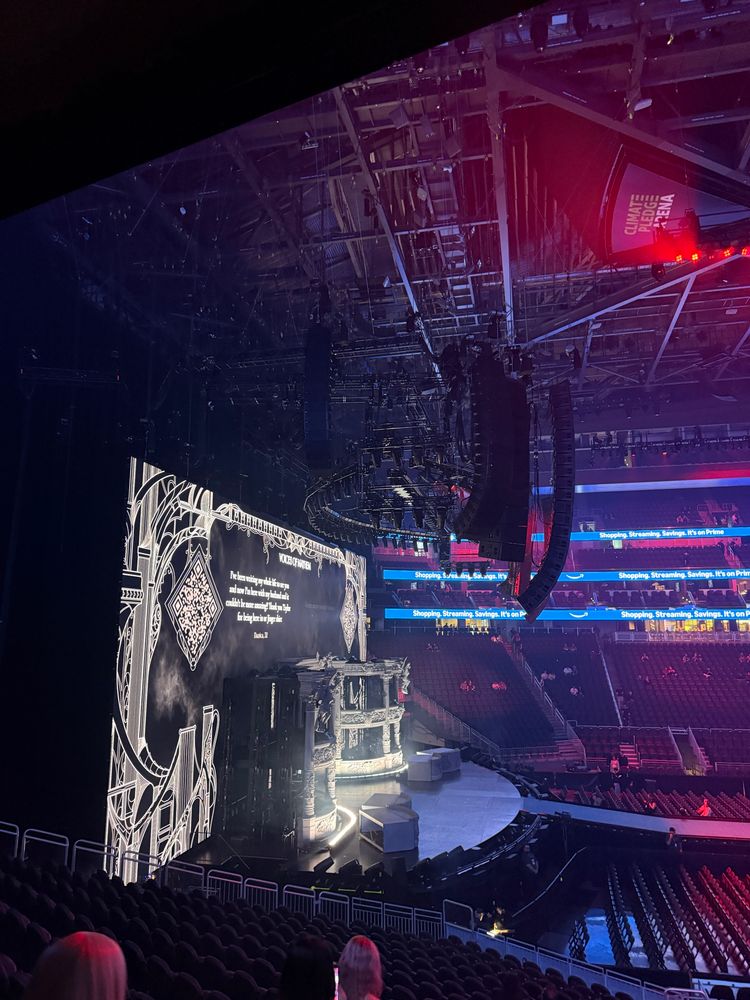 A large indoor arena, Climate Pledge Arena, is shown preparing for a concert or performance. The stage features ornate gothic-style architecture with dramatic lighting, a curved front, and props. A large digital screen displays stylized text and QR codes framed by intricate black-and-white designs. The audience seating is mostly empty, with a few people scattered around, suggesting the event has not yet started. Overhead rigging and lights are visible above the stage. LED ribbon displays around the arena advertise Amazon Prime.