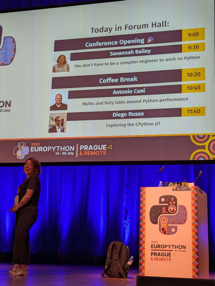 Me standing in front of a large screen with a list of talks for the day before the day. I’m listed as the opening keynote. I look happy and excited!