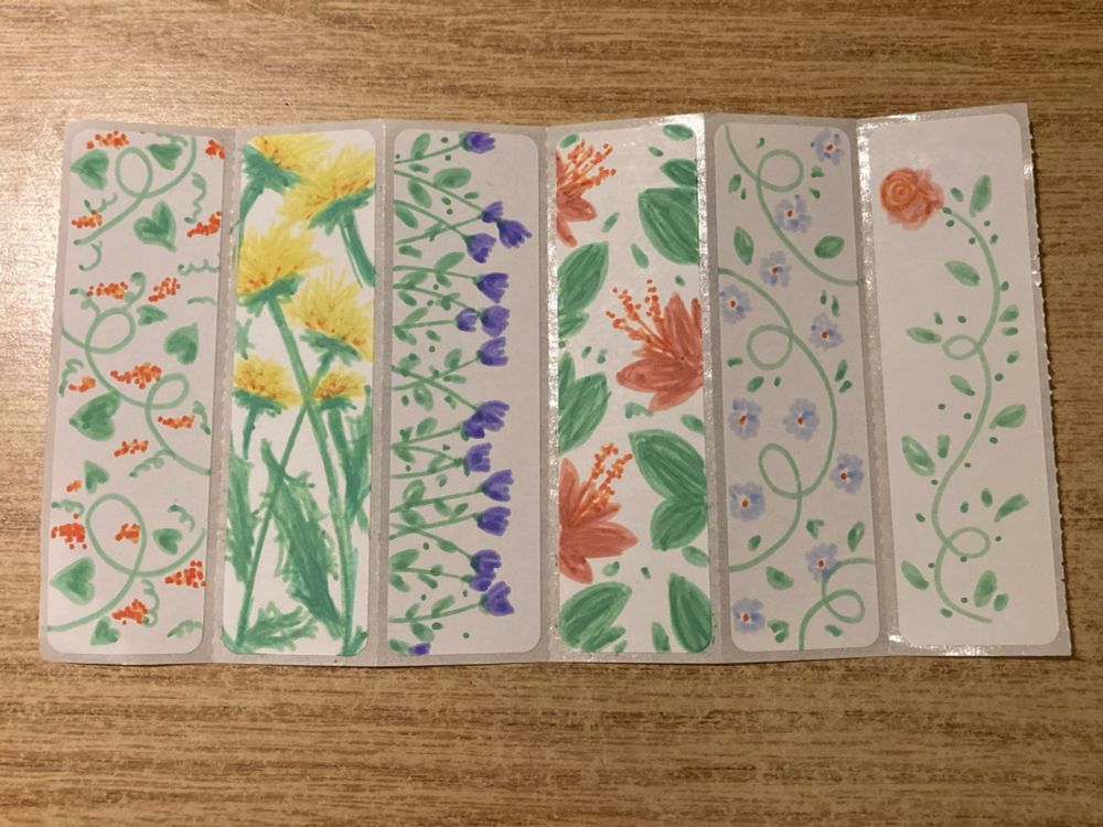 Six thermal print labels decorated with hand drawn pictures of flowers. From left to right: vines with small orange clusters, dandelions, purple tulips, red hibiscus, a small vine with small blue flowers and finally, a rose with a long twisting stem.
