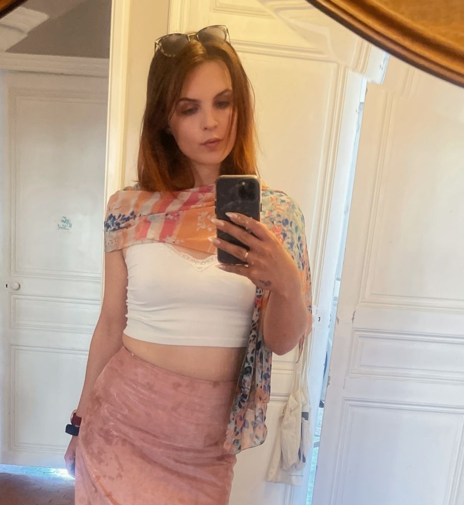 mirror selfie of a red head women with a scarf around her neck a white crop tank and a pink dress. there are sun glasses on her head and her nails are done