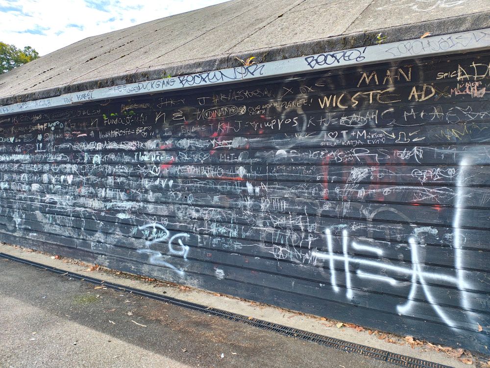 Wall with dark wooden slats at the edge of a playground, covered in graffiti by numerous hands in white, yellow and red spray paint.